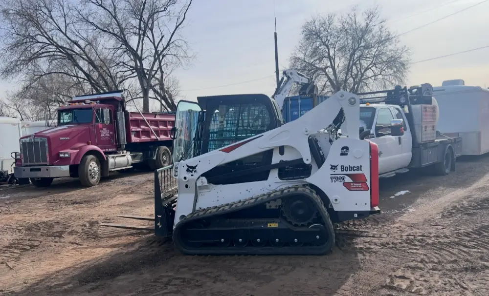 A Bobcat skid steer loader parked amidst a construction site, with a pink dump truck and pickup truck visible in the background.