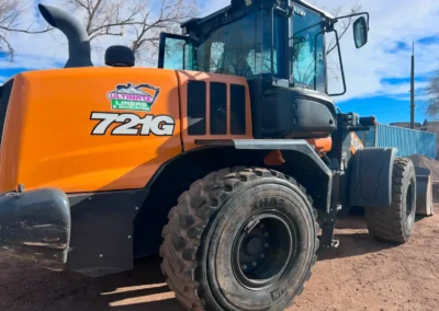 An orange heavy machinery loader, model 721G, with a 'Ultimate Liners & Excavation' sticker, parked on gravel under a blue sky.