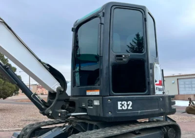 A close-up of a Bobcat E32 excavator with a black cab and white arm, set against a cloudy sky and industrial background.