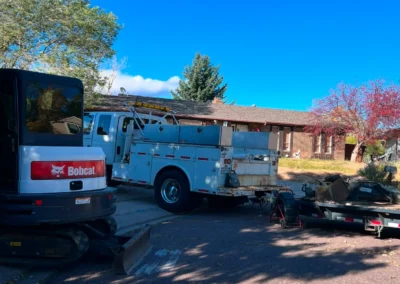 A Bobcat vehicle and a utility truck are parked on a residential street, with a trailer holding construction materials in the background.